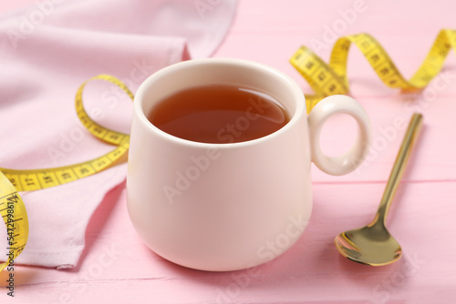 Cup of herbal diet tea, spoon and measuring tape on pink wooden table, closeup. Weight loss concept