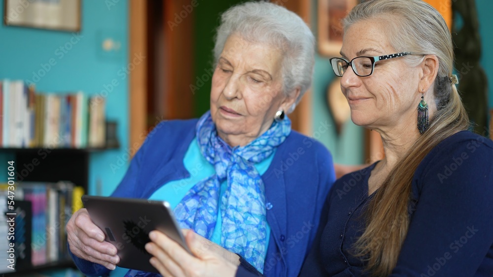 Happy elderly senior woman and mature woman look at tablet computer while drinking tea on a couch in a living room. Happy elderly senior woman and mature woman look at tablet computer while drinking tea on a couch in a living room.
