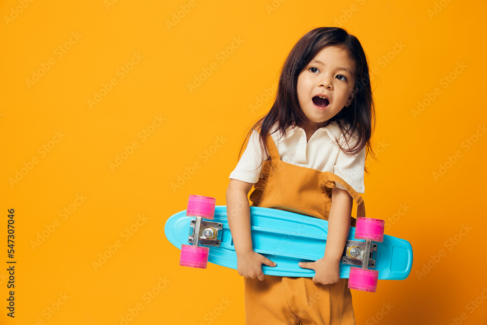 beautiful, little girl of preschool age, stands on an background in a sundress and holds a light blue skateboard in her hands, smiling broadly with delight. Photo with space for an advertising layout beautiful, little girl of preschool age, stands on an background in a sundress and holds a light blue skateboard in her hands, smiling broadly with delight. Photo with space for an advertising layout