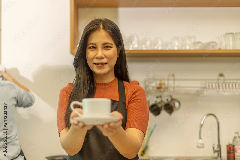 Young female coffee shop owner is holding a cup of hot coffee to customer with care. Lady with warm hospitality and care hands a cup of hot drink to her client in a small shop. Young female coffee shop owner is holding a cup of hot coffee to customer with care. Lady with warm hospitality and care hands a cup of hot drink to her client in a small shop.