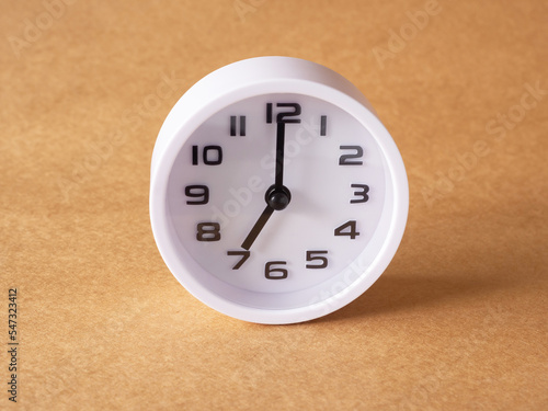 Close up, white clock shows time at 7 o'clock on the office brown workplace tabletop. Copy space for your text. Business and education concept..