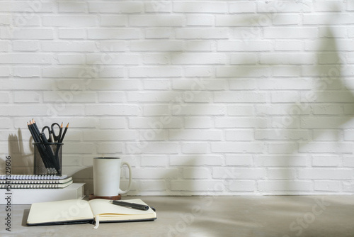 Comfortable workplace with coffee cup  notebook and pencil holder on white table against brick wall. Copy space for your text.