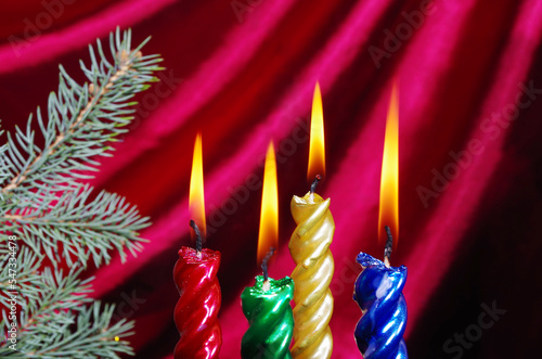 Four burning Christmas candles and a decorated fir tree against the background of a burgundy curtain. 