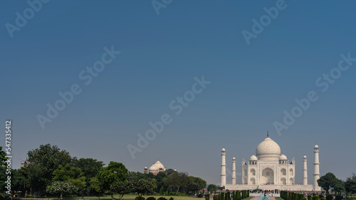 Beautiful ancient mausoleum of the Taj Mahal against the blue sky. Minarets, towers, domes, arches made of white marble. Green vegetation in the park. A lot of people are sightseeing. Copy space.