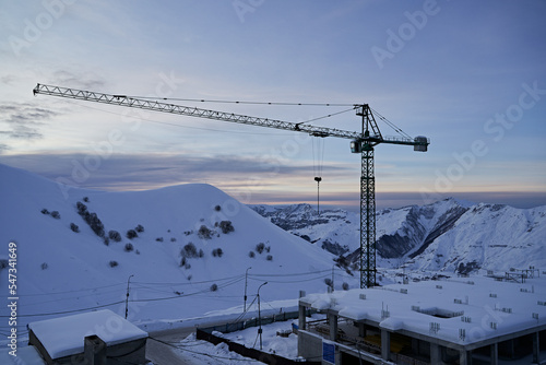 Construction site of ski resort hotel chalet framework and crane with snowy mountain ridge on winter sunrise on background. Mountains range covered with snow powder against the residental building.