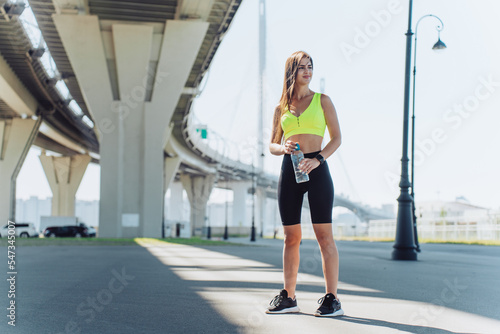 Attractive brunette hispanic girl in sportswear standing outside under viaduct smiles happy to be fit, active and healthy young woman. Beautiful successful female at workout holds bottle of water.