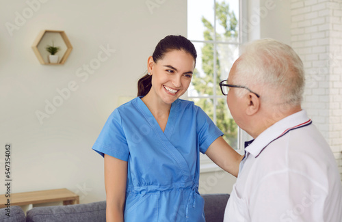 Friendly brunette young nurse smiling young woman is supporting helping old man in hospital. She is touching patient on shoulder and looking at him. Support, help for old patiens in clinic concept.
