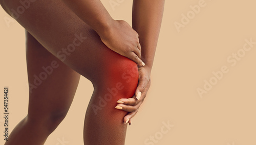 Knee pain as result of injuries such ruptured ligaments  ruptured cartilage or improper exercise. Close up of woman holding sore spot on her knee which is highlighted by red light. Beige background.