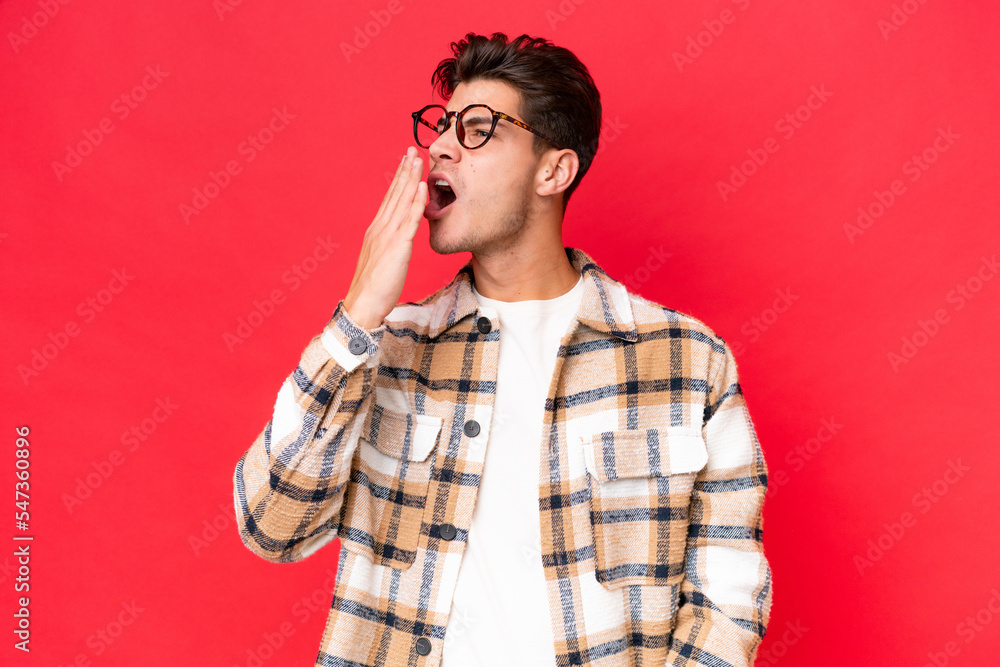 Young caucasian handsome man isolated on red background yawning and covering wide open mouth with hand Young caucasian handsome man isolated on red background yawning and covering wide open mouth with hand