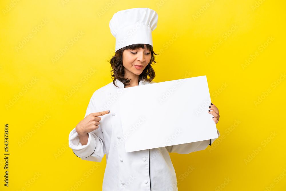 Young chef latin woman isolated on yellow background holding an empty placard with happy expression and pointing it Young chef latin woman isolated on yellow background holding an empty placard with happy expression and pointing it
