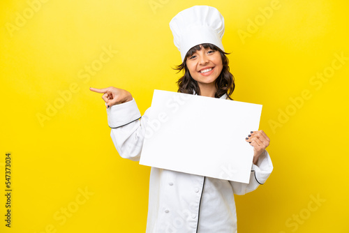 Young chef latin woman isolated on yellow background holding an empty placard and pointing side photo