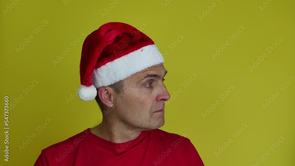 A man in a Santa hat in a red T-shirt on a yellow background turns his head to the left and right. Christmas concept. A man in a Santa hat in a red T-shirt on a yellow background turns his head to the left and right. Christmas concept.