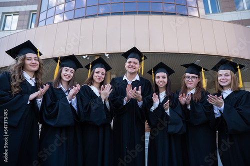 Seven graduates in robes stand in a row and clap outdoors.