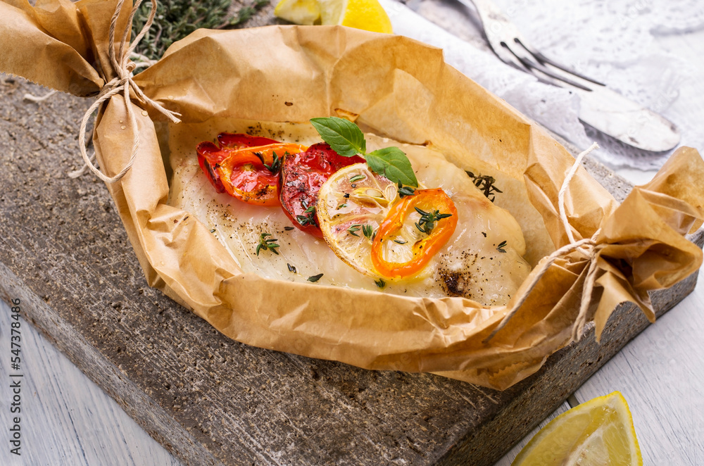 Traditional baked cod fish fillet with vegetable and herbs in backing paper served as close-up on a Nordic design plate