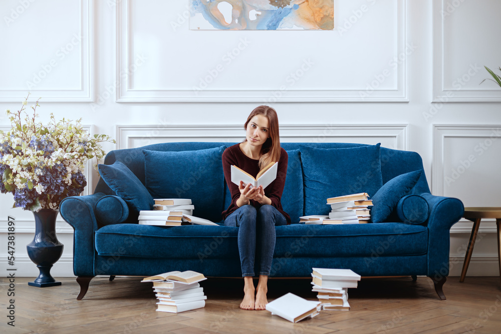 Young woman among stacks of books on a spacious sofa in the living room.