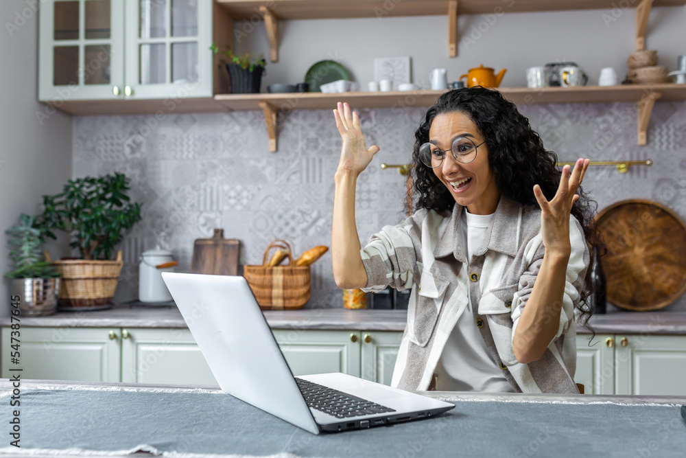 Positive and creative Latin American woman sitting at home at the kitchen table and working, studying at the laptop. He talks to the camera, explains with his hands, smiles Positive and creative Latin American woman sitting at home at the kitchen table and working, studying at the laptop. He talks to the camera, explains with his hands, smiles