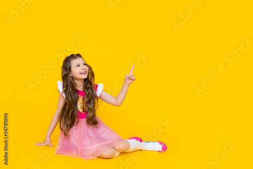 A little lady sits on a yellow isolated background and points her finger at your advertisement on a yellow isolated background. A beautiful girl with long hair in a pink dress is smiling broadly.