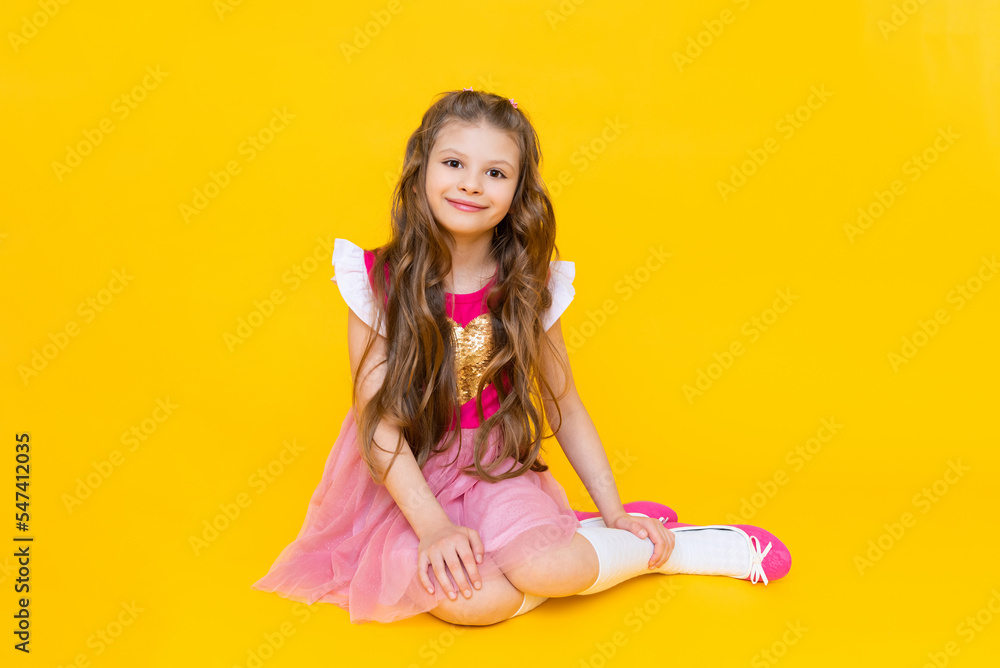 The little lady is sitting on a yellow isolated background. A beautiful girl with long hair in a pink dress is smiling broadly. The little lady is sitting on a yellow isolated background. A beautiful girl with long hair in a pink dress is smiling broadly.