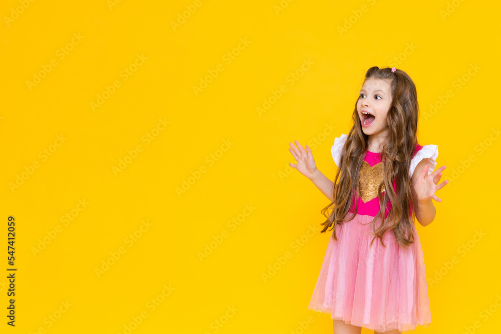A little girl points to your ad on a yellow isolated background. A beautiful child in a pink dress points to an empty place and smiles broadly.