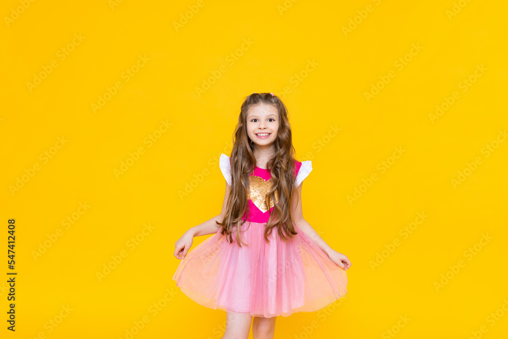 A little princess in a pink dress with curly hair is smiling broadly. A beautiful, charming child on a yellow isolated background. A little princess in a pink dress with curly hair is smiling broadly. A beautiful, charming child on a yellow isolated background.