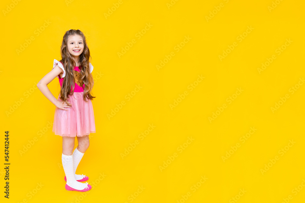 A beautiful little girl in full height in a pink dress on a yellow isolated background. Happy little princess with long curly hair.