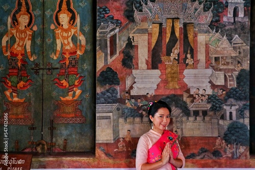 A Thai lady in Mon costume smiles and put hands together in salute at the mural wall of temple