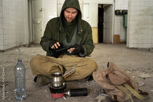 фотографія a dirty tired man sits on the floor in an abandoned room, warms her hands over a gas burner and prepares dinner for herself