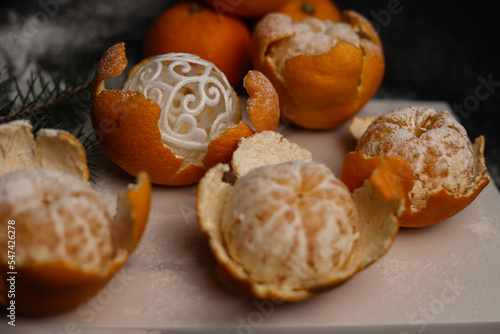 sweet orange juicy tangerines in a peeled peel lie on a black background and a black kitchen tray and in one peel a New Year's ball next to a spruce Christmas branch and sprinkled with white powder.fo