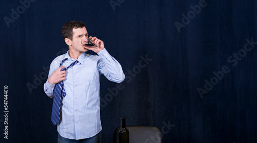 White man in business shirt stands against dark background and drinks red wine from glass with large gulps. He rips his tie off neck with hand. Stressful situation.