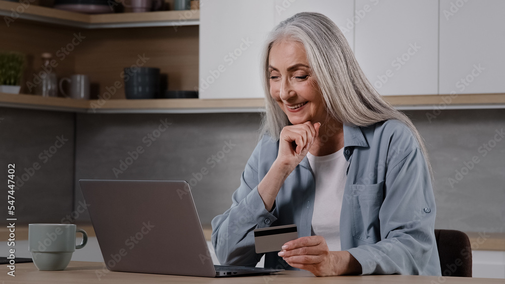 Happy old female caucasian consumer grey-haired granny shopper pay internet order on laptop sits in kitchen hold banking card use online banking app donating money deposit rejoice approved credit loan Happy old female caucasian consumer grey-haired granny shopper pay internet order on laptop sits in kitchen hold banking card use online banking app donating money deposit rejoice approved credit loan