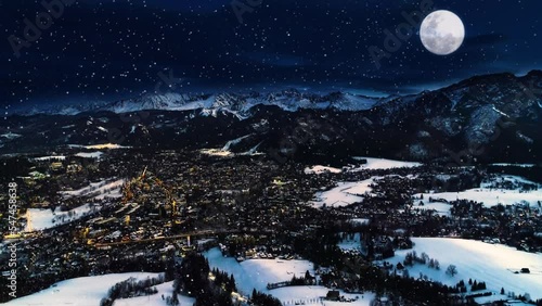 Snowy winter night, epic aerial view. Magical winter in the mountain village. Illuminated alpine city covered with snow. White mountains, ski resorts and big shining moon. Christmasy snowy landscape.
