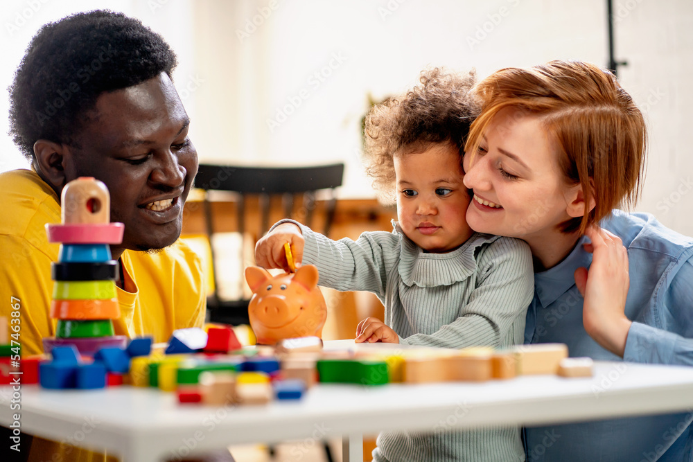 A child is putting a coin into a piggy- bank. Savings bank accounts for kids concept. Child saving money for future - concepts. Multi-ethnic  family