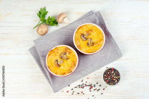 Chicken and mushroom casserole with golden crust, julienne with baked cheese, creamy gratin in portion ramekin clay pot on white wooden background. French cuisine. Top view, flat lay