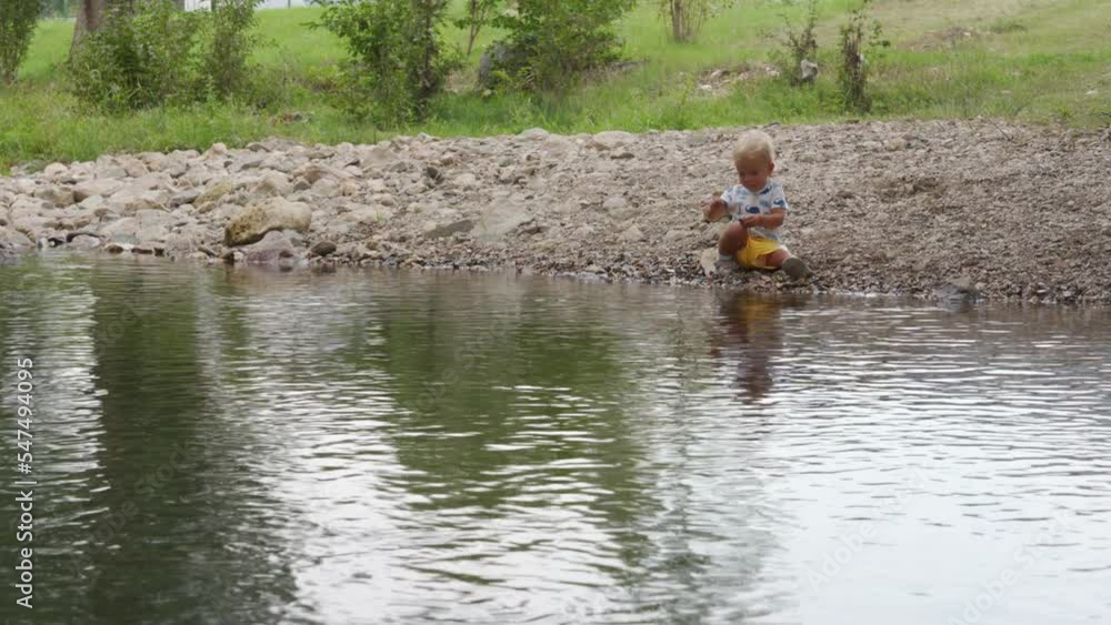 Little child playing on river bank in a summer park, 1.5 year old toddler enjoys throwing pebbles into the water. Little child playing on river bank in a summer park, 1.5 year old toddler enjoys throwing pebbles into the water.