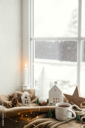 Warm cup of tea, christmas decorations, lights, little house, star on cozy blanket on windowsill. Winter hygge, Christmas still life. Cozy home on snowy day. Atmospheric scandinavian mood