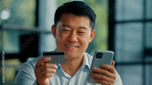 Happy Asian man client buyer businessman use banking mobile phone app at office hold plastic credit card enter secure code buy goods in internet store shopping online cryptocurrency money transaction 