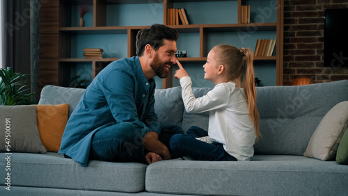 Playful caucasian young family daddy and small daughter sitting comfortable on couch loving cute girl touching father nose funny joke playing game with parent having fun spending free time together photo