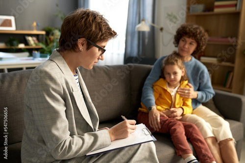Social worker making notes in document while talking to difficult child and his mom at home photo