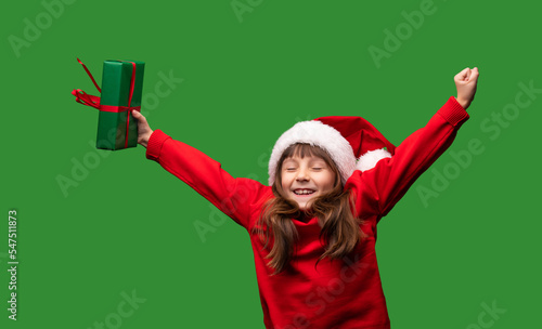 Wow, holiday. A girl in a Santa hat is very emotionally happy with gifts for Christmas and New Year 2023. Happy childhood. Portrait of a child isolated on a green background