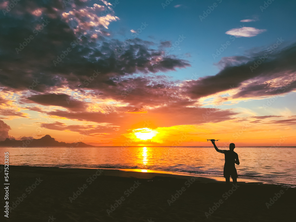 A man shooting sunset by drone. The silhouette of an operator holding quadracopter in hands, watching bright orange sunset over sea, cloudy sky. Beautiful nature landscape. Popular tourist destination
