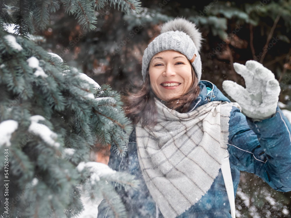 Smiling woman in cable knitted hat is playing with snow. Fun in park between snowy fir trees. Woman laughs as she is throwing snowball. Cold season. Smiling woman in cable knitted hat is playing with snow. Fun in park between snowy fir trees. Woman laughs as she is throwing snowball. Cold season.