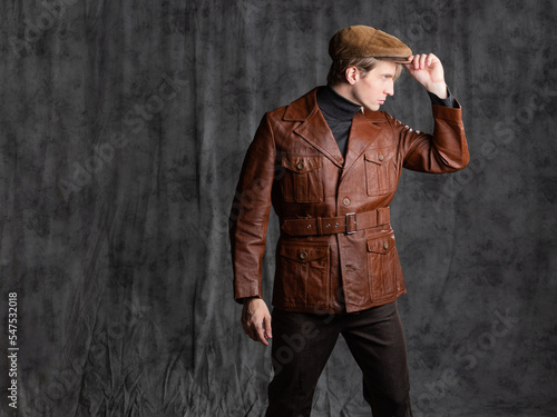 young man, a retro secret agent, a guy in a brown leather jacket and a flat cap with a gun in his hand. Posing in the studio on a gray background