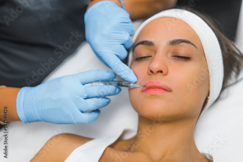 Aesthetic medicine concept. Lip filler procedure closeup. Attractive young adult caucasian woman portrait. Professional lip injection done by unrecognizable expert in blue protective gloves. High
