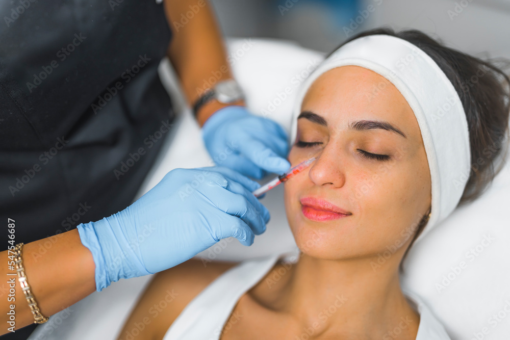 Fototapeta premium Portrait of an attractive caucasian European woman in her 30s wearing white protective hairband during collagen injection procedure done by a professional beautician in protective gloves. Aesthetic
