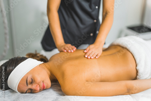Taking care of one's health through SPA body care. Satisfied happy caucasian young woman with hairband above her forehead during relaxing back massage performed by unrecognizable massage therapist in