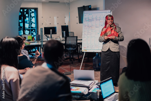 An older Muslim businesswoman presents a project to a young diverse team of people in a modern office