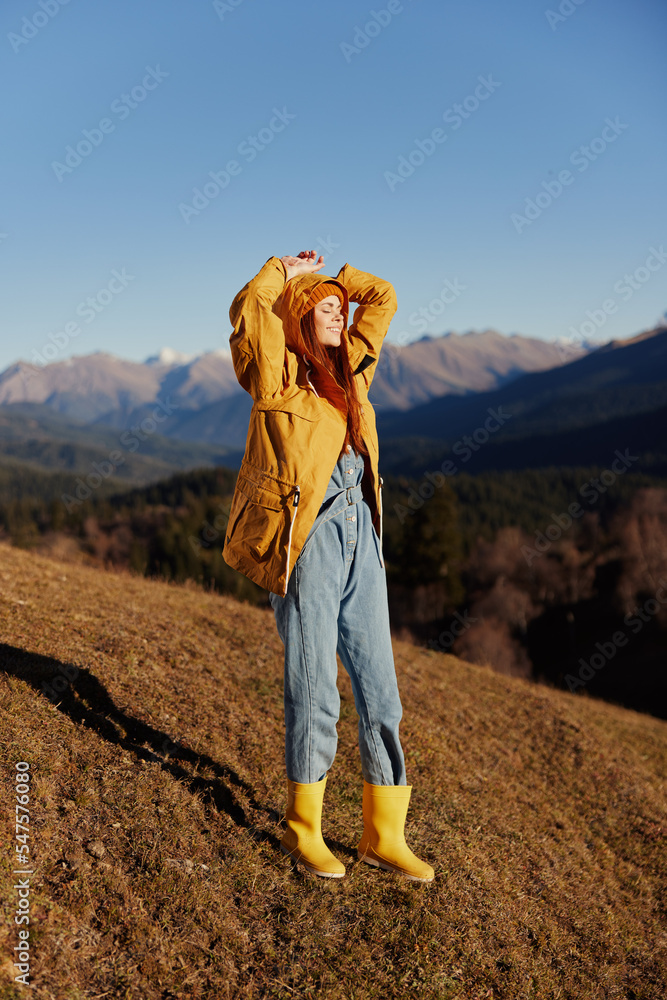 Woman autumn smile with teeth full-length hands up happiness walking on the hill and looking at the mountains in a yellow raincoat and jeans happy trip in the sun sunset hiking, freedom lifestyle 
