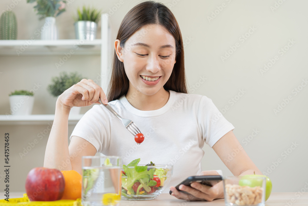 Dieting, smile asian young woman eating, holding fork at tomato, diet plan nutrition with fresh vegetables salad, enjoy meal while using smartphone. Nutritionist of healthy, nutrition of weight loss. Dieting, smile asian young woman eating, holding fork at tomato, diet plan nutrition with fresh vegetables salad, enjoy meal while using smartphone. Nutritionist of healthy, nutrition of weight loss.