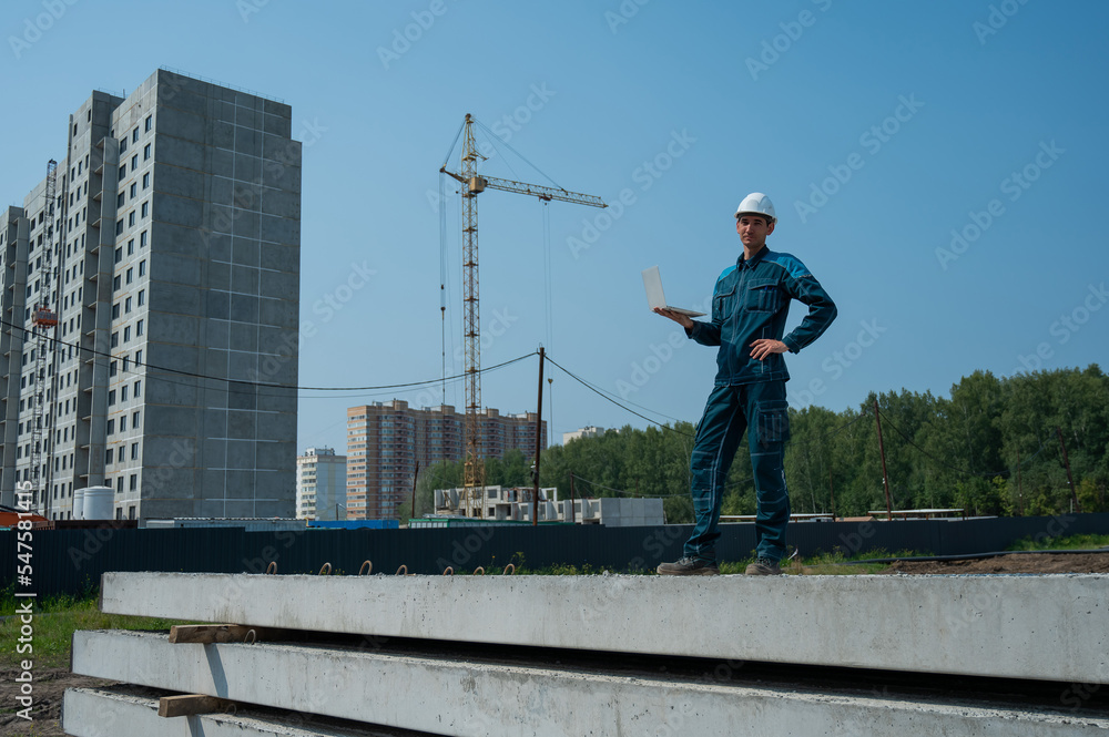 A male builder in a helmet stands against the background of a construction site with a laptop in his hands. A male builder in a helmet stands against the background of a construction site with a laptop in his hands.