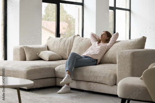 Young attractive calm woman in casual clothes spend free time relaxing on comfy sofa with eyes closed. Housewife finished housework chores, resting seated on couch in fashionable living room. Leisure